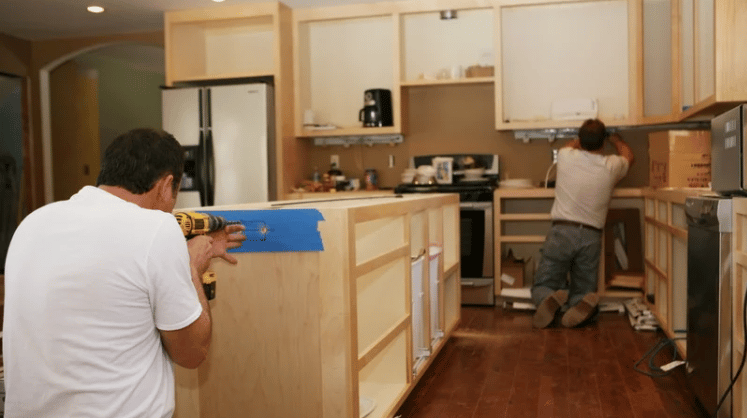Kitchen under renovation with two workers installing unfinished wooden cabinets.