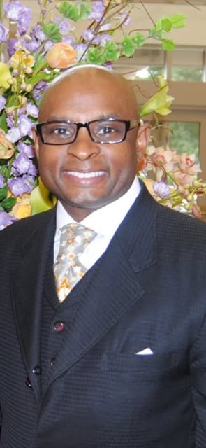 Dwain Byrum wearing glasses and a dark suit with a patterned tie, posed before flowers.
