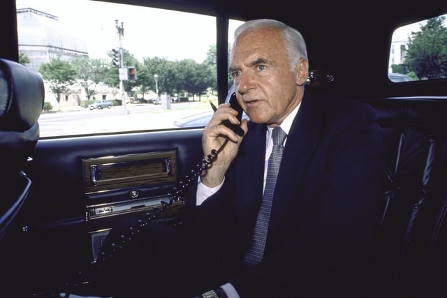 Older man in a suit talking on a corded telephone inside a car.