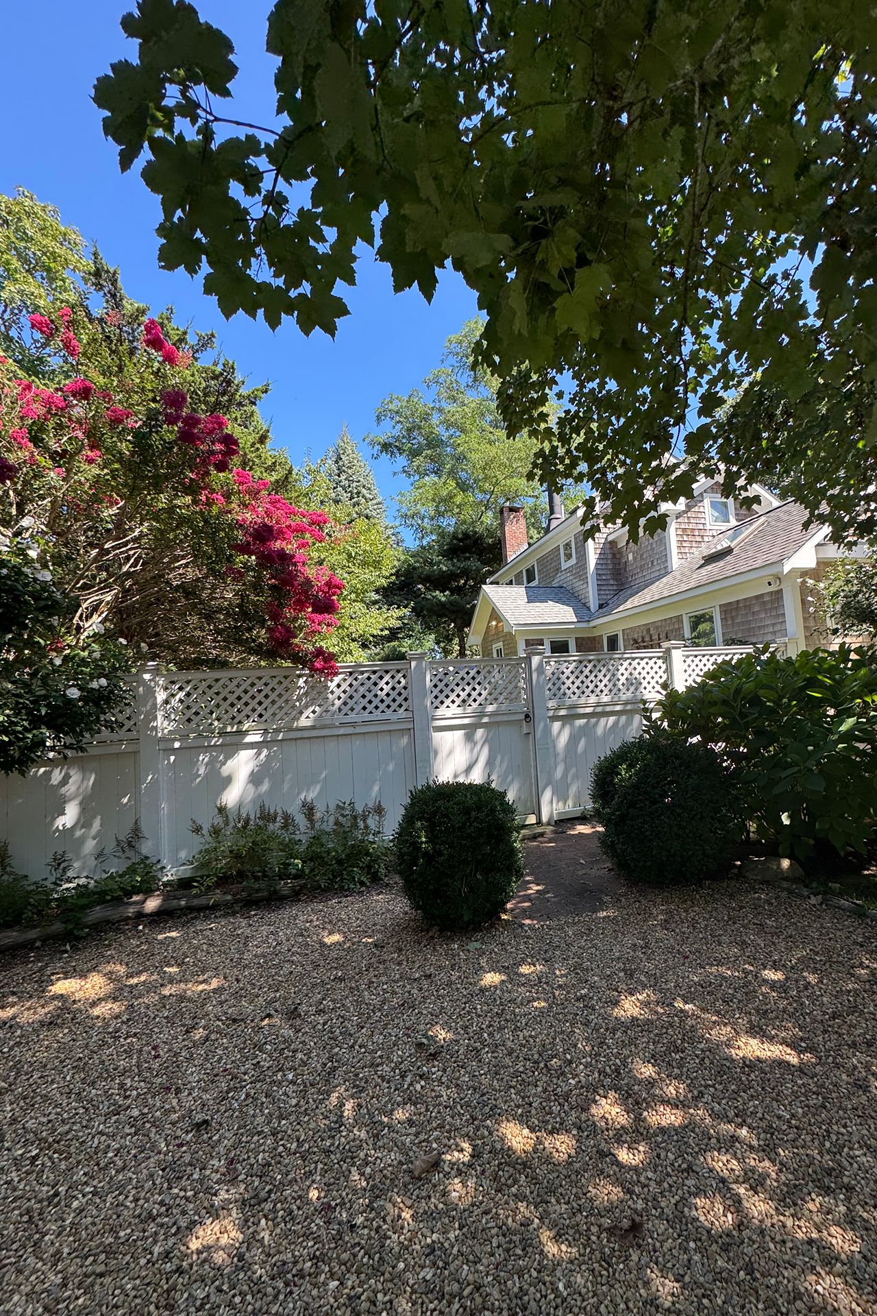 Shingle-sided home behind a white lattice fence, framed by flowering shrubs and a gravel courtyard.