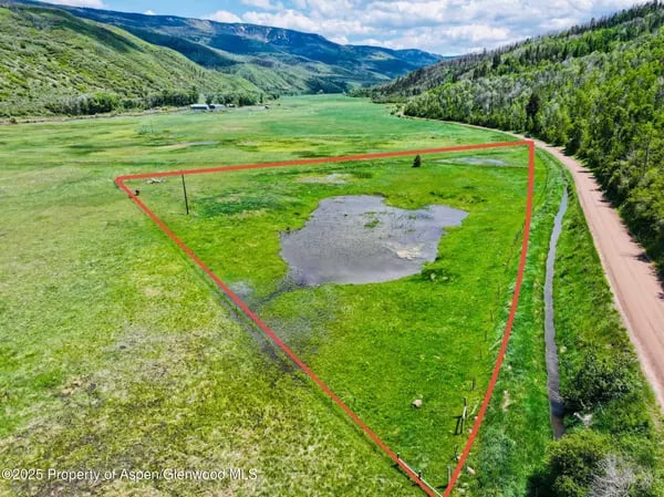 Vacant green parcel outlined in red, with a wet area and dirt road beside forested mountains.