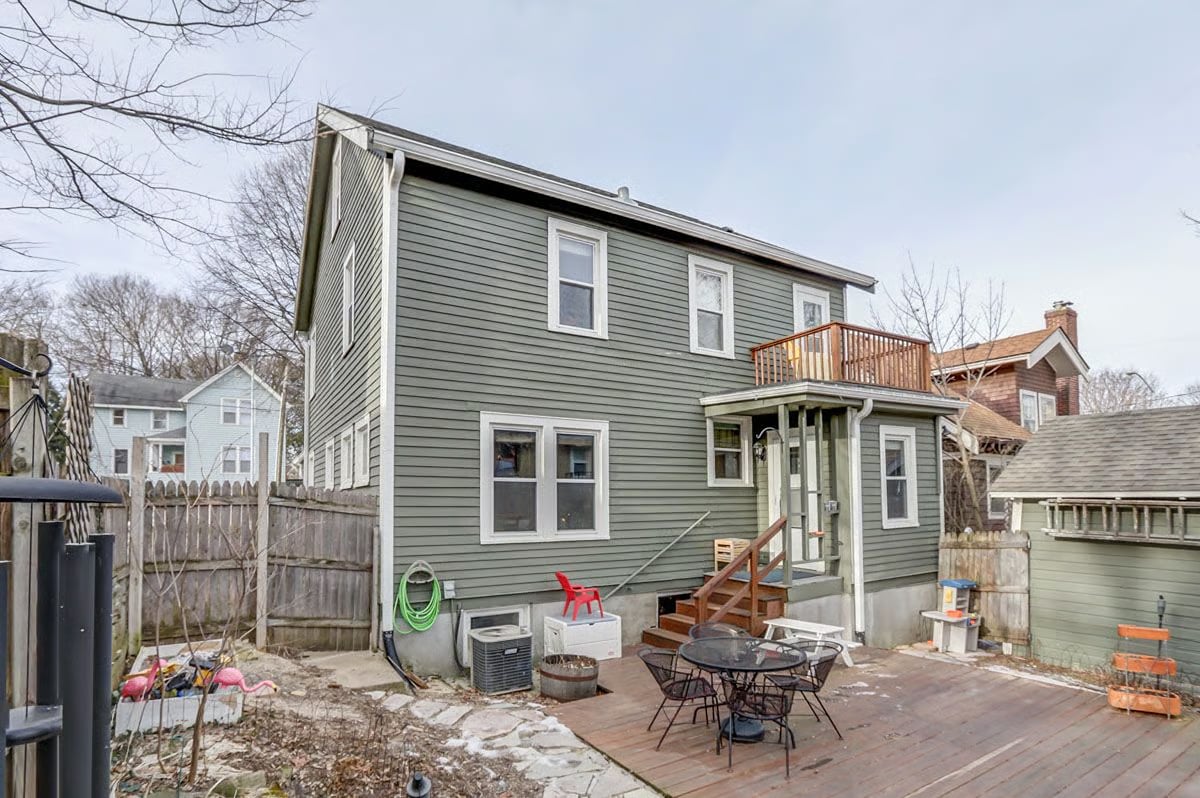 Two-story sage-green house with a small balcony and patio dining area.