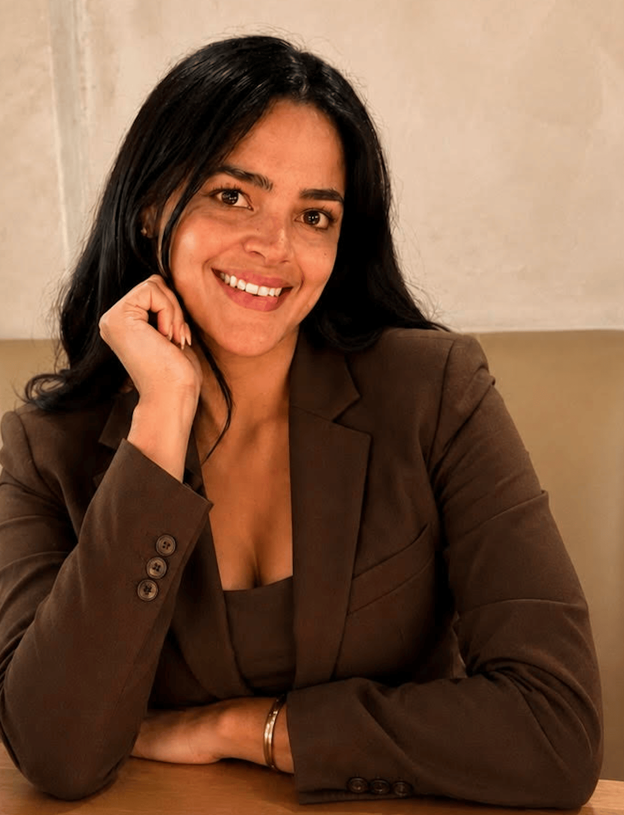 Smiling woman in a brown blazer, resting her hand against her cheek.