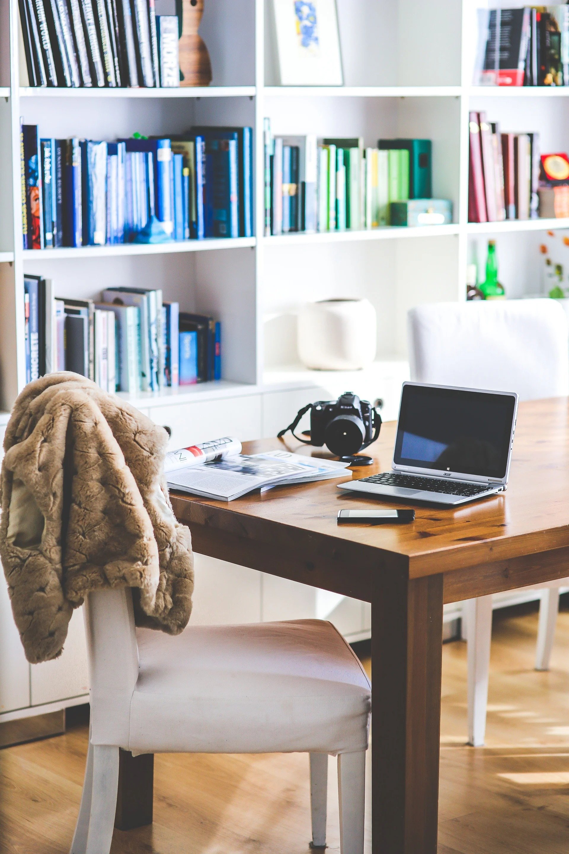 Furnished home office with a wooden desk, laptop, camera, and white built-in bookshelves.