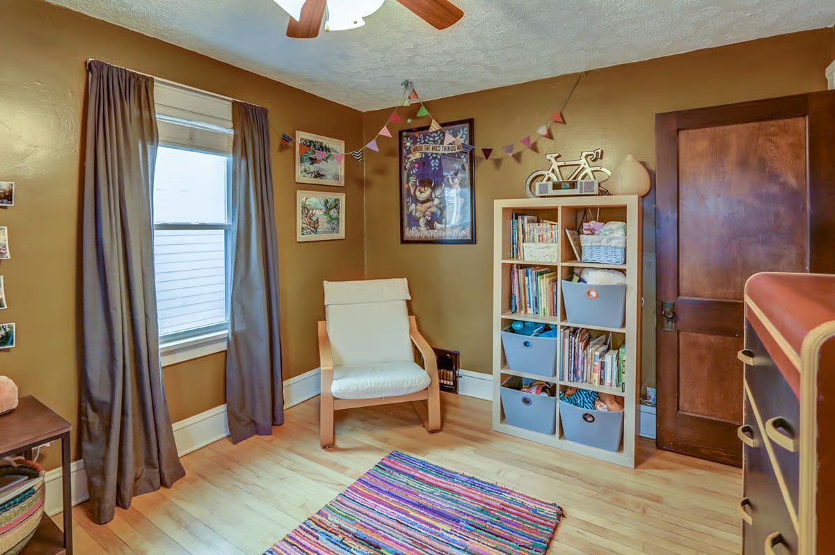Bedroom with ochre walls, a white chair, bookcase, and colorful striped rug.