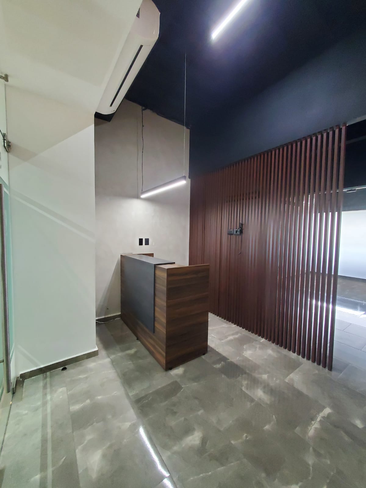 Office reception area with a wood desk, slatted partition, and polished stone flooring.