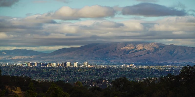 Panoramic view of Silicon Valley with downtown buildings in the distance and rolling hills under a partly cloudy sky.