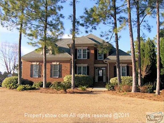 Two-story brick house framed by tall pine trees across a broad lawn.