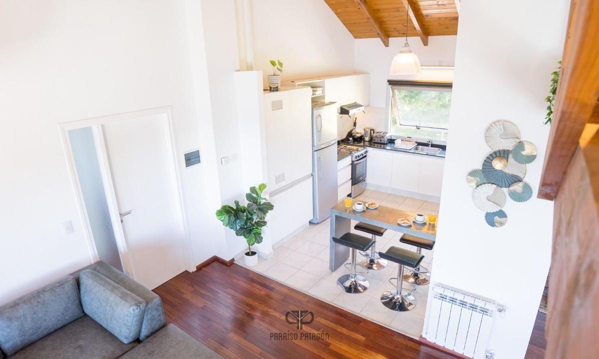 Open-plan apartment kitchen and living area with a breakfast island, gray sofa, and exposed timber ceiling.