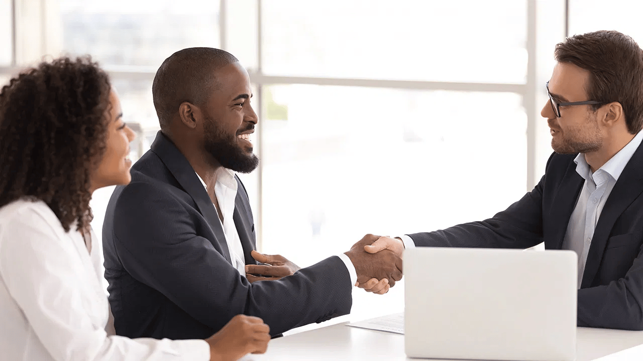 Two men shake hands across a table as a woman watches in a bright office.