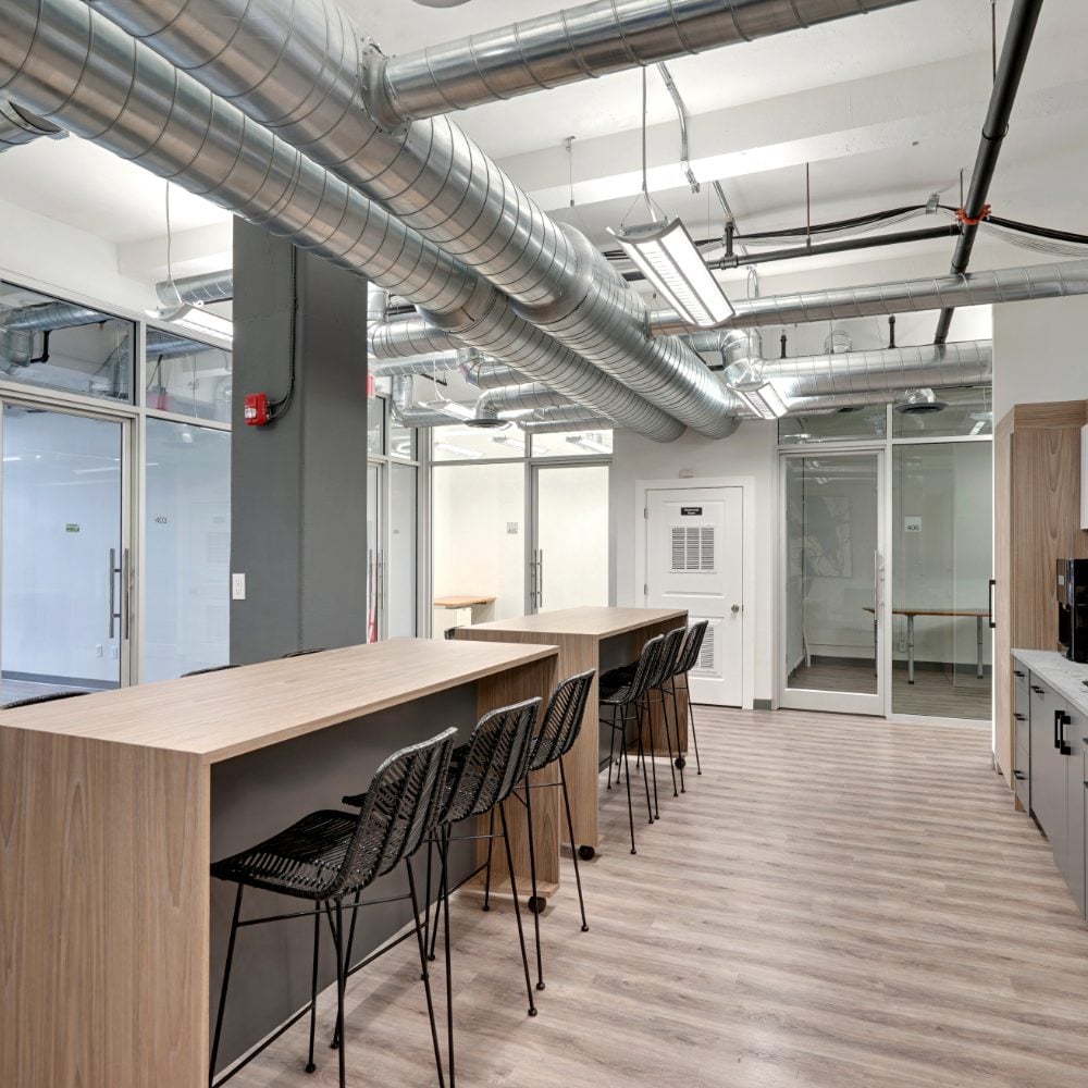 Modern office with a long wood counter, black stools, glass partitions, and exposed ventilation ducts.