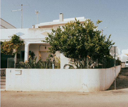 White corner-lot house partly hidden by a leafy tree behind a low wall.