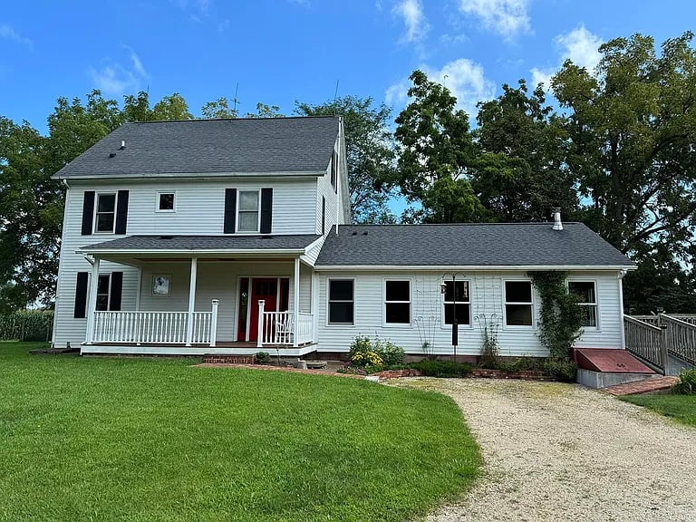 White two-story house with a covered porch, red front door, and gravel driveway.