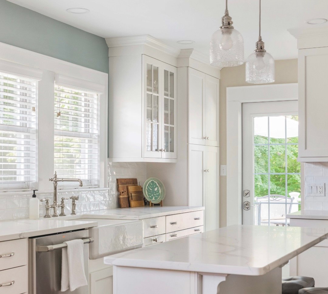 Bright white kitchen with a farmhouse sink, marble island, glass-front cabinets, and garden views.