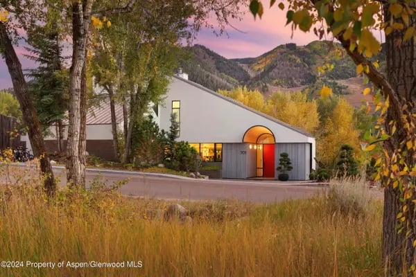 Modern angular home with a bright red entrance framed by autumn trees, with mountains behind it.