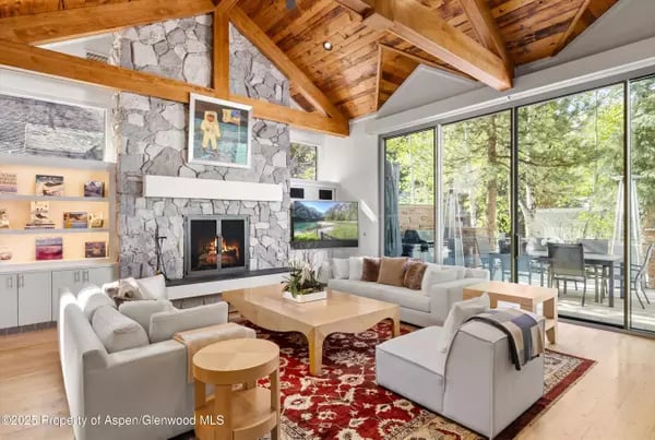 Living room with a vaulted timber ceiling, stone fireplace, and large windows overlooking trees.