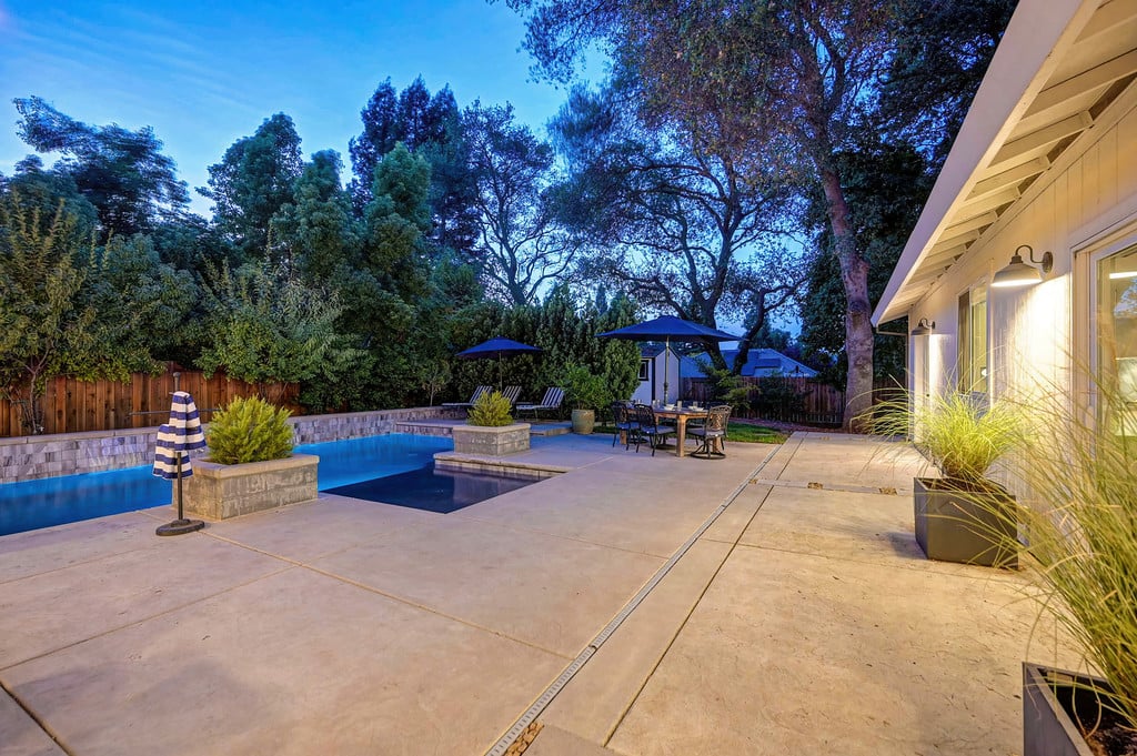 Backyard pool and patio at dusk beside the home, with outdoor dining furniture and mature trees.