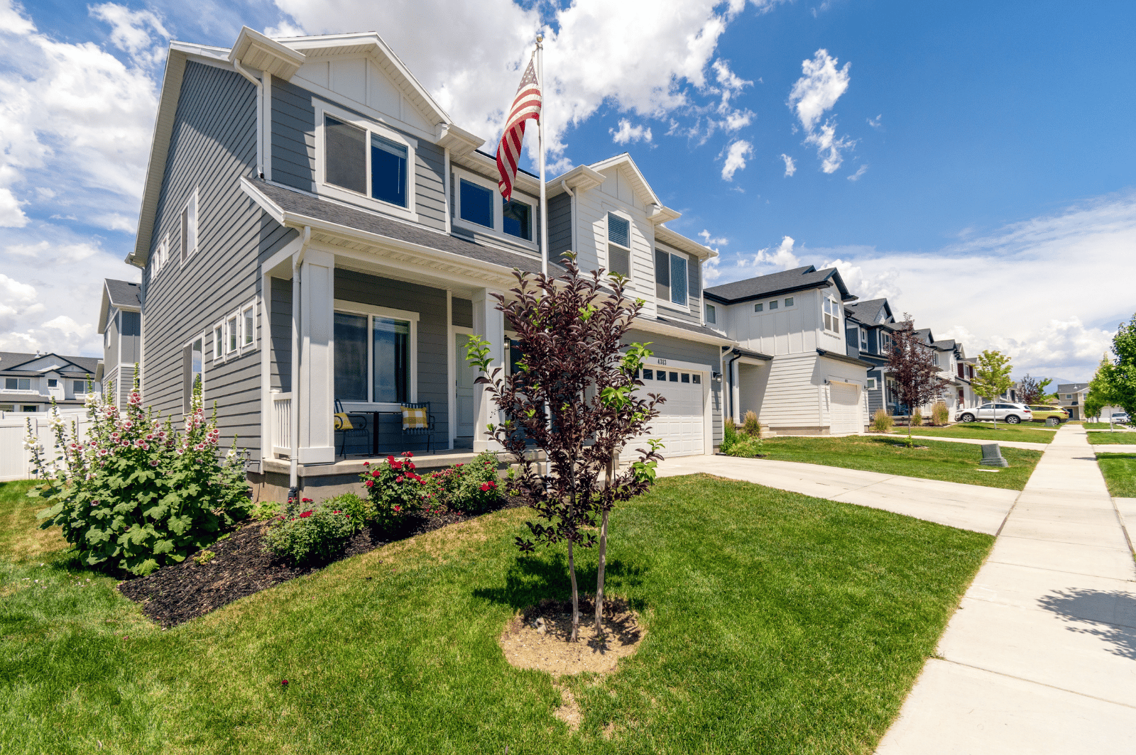 Two-story gray home with a covered porch, American flag, flower beds, and front lawn.