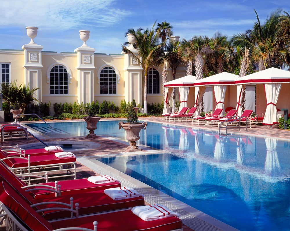 Resort pool with red loungers, white cabanas, palm trees, and an ornate building.