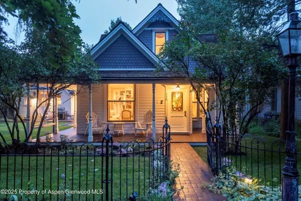 Two-story home with a warmly lit porch, garden path, and iron fence at dusk.