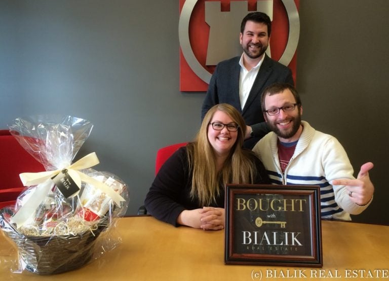 Steven and Katie celebrate their home purchase with a Bialik Real Estate agent and a framed sign.