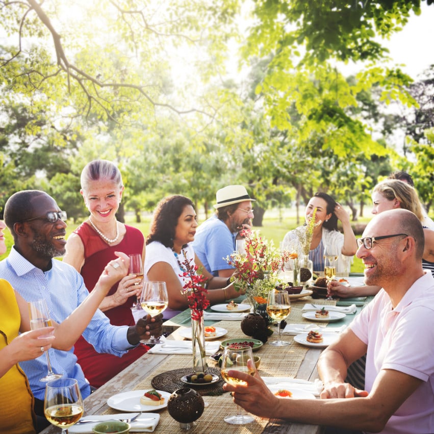 Adults sharing a meal at a long outdoor table beneath leafy trees.