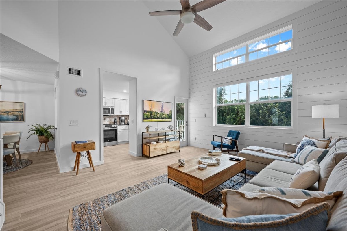 Furnished condominium living room with vaulted ceiling, large windows, and an open kitchen.
