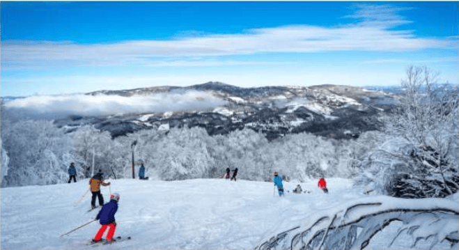 Skiers descend a snowy slope beneath frosted trees and distant mountain ridges.
