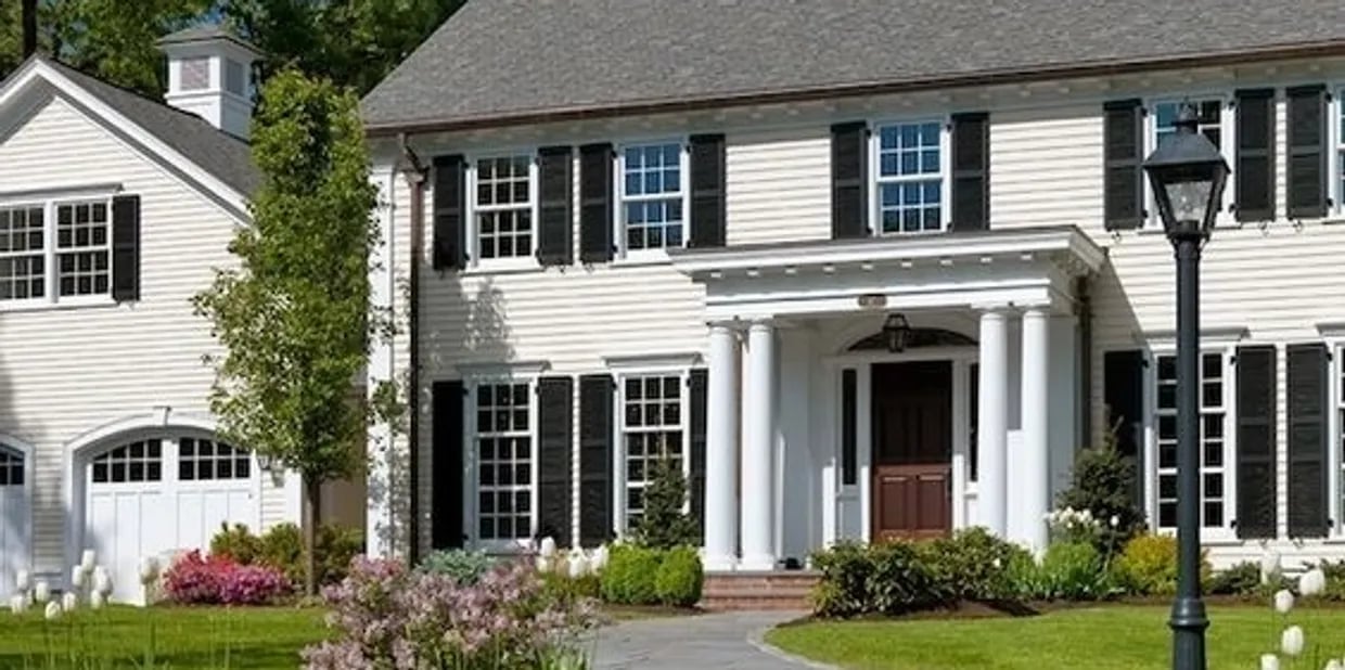 White Colonial home with black shutters, a columned entrance, and landscaped flower beds.