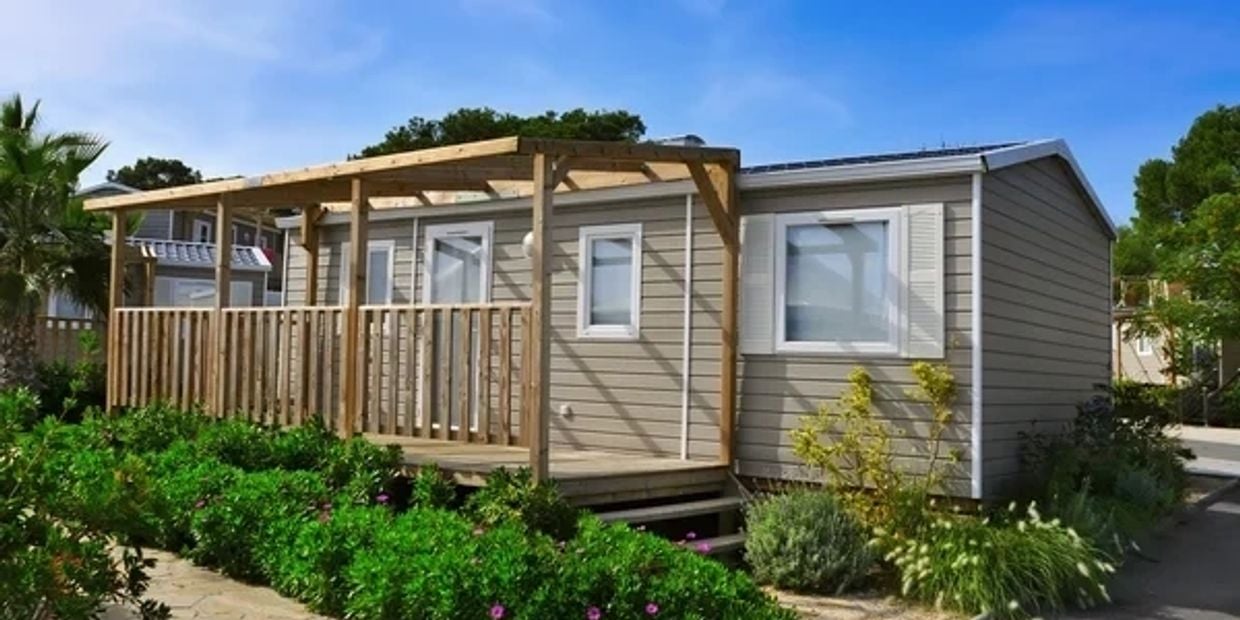 Gray mobile home with a wooden porch and pergola, surrounded by landscaped shrubs.