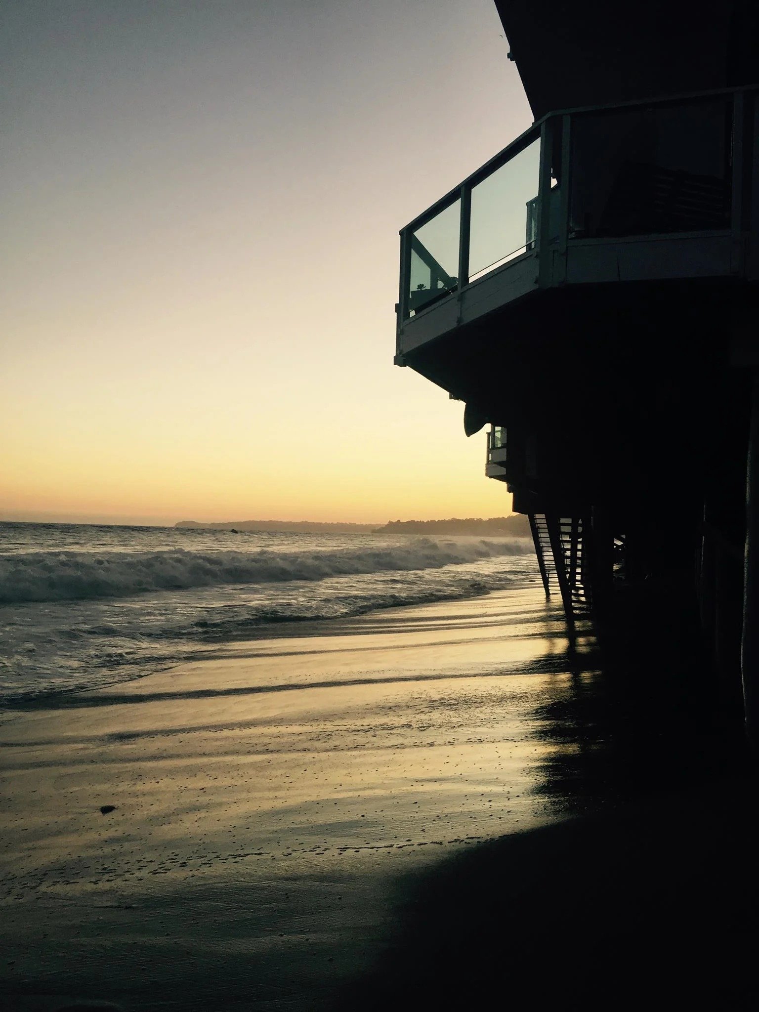 Sunset waves reach the shore beneath a silhouetted beachfront building on stilts.