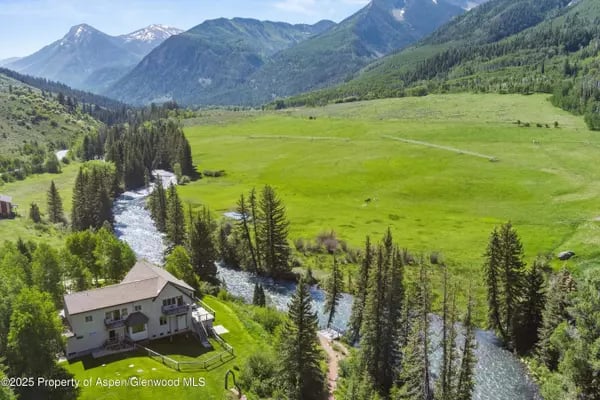 Aerial view of a riverside home beside a green meadow, with forested mountains beyond.