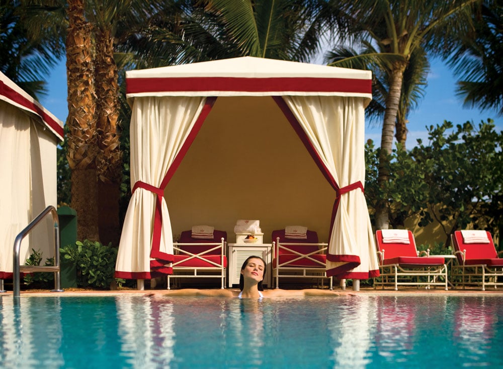 Resort swimming pool with a curtained cabana, red loungers, and a swimmer.