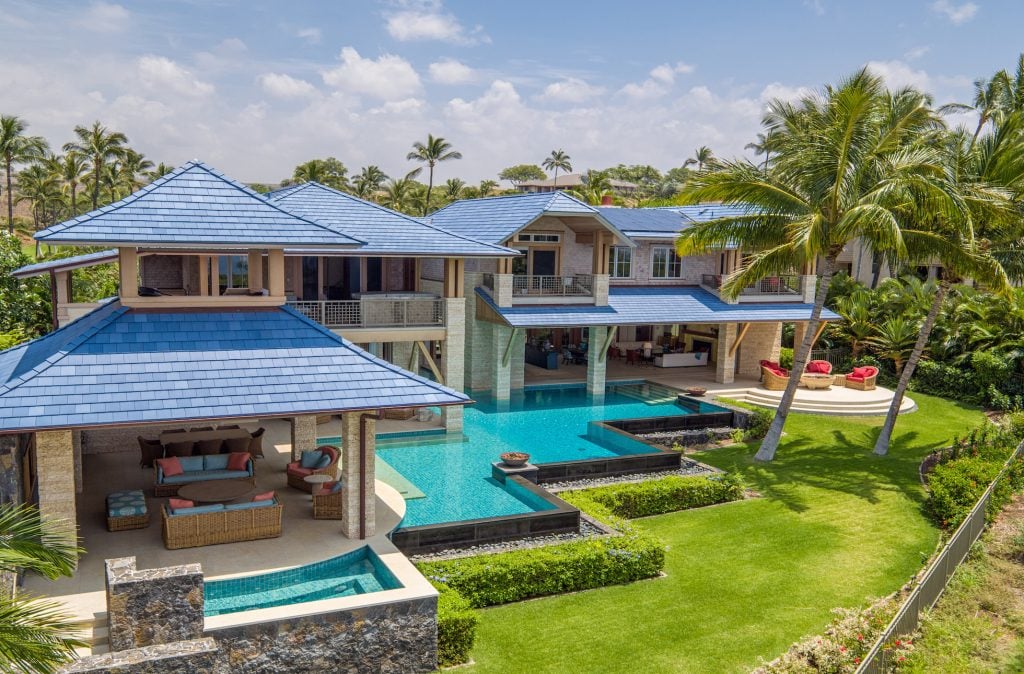 Tropical luxury home with blue tiled roofs, a multi-level pool, and palm-lined lawn.