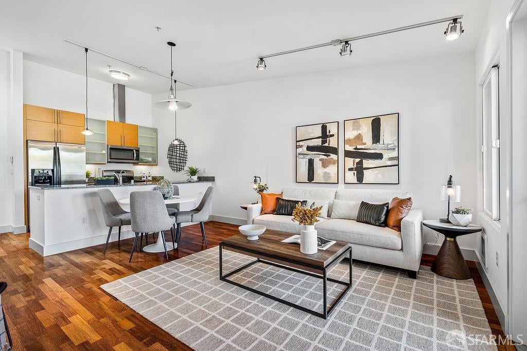 Open-plan living room and kitchen with warm wood floors, contemporary furnishings, and a gray geometric rug.