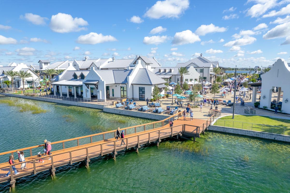Waterfront village with white-roofed buildings, outdoor gathering spaces, and a boardwalk over the water.