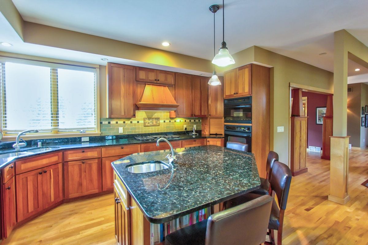 Kitchen with warm wood cabinetry, dark granite island, built-in ovens, and hardwood floors.