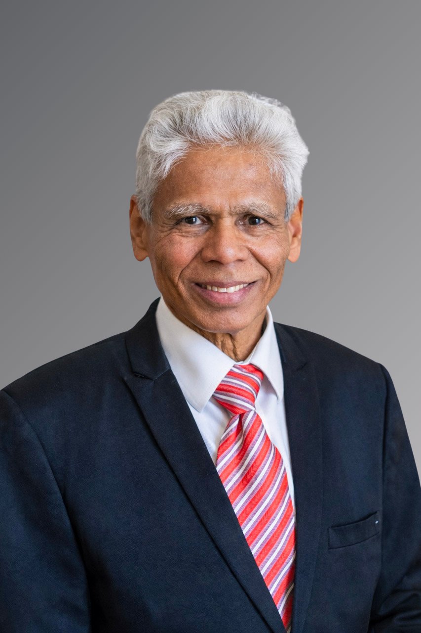 Suresh Sharma in a dark suit and red striped tie, smiling against a gray background.
