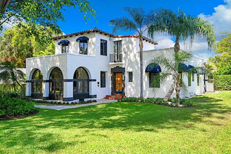 White two-story stucco home with arched openings, palm trees, and a broad green lawn.