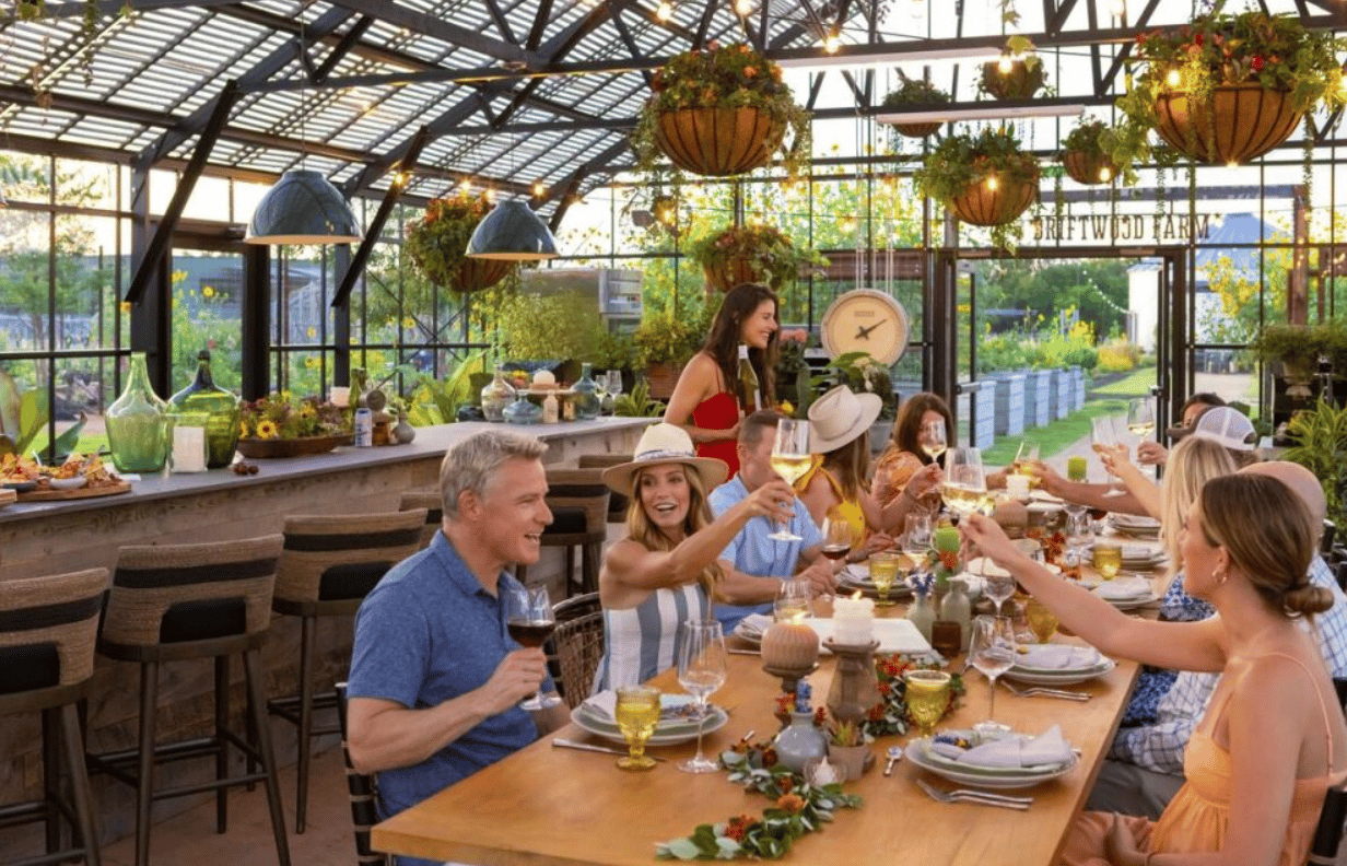 Guests toast around a long table beneath hanging planters in a glass-roofed restaurant.