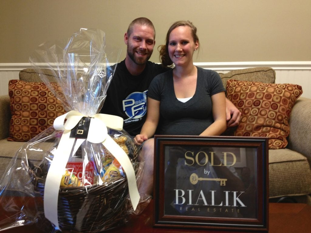 A couple celebrates a home closing beside a gift basket and Bialik Real Estate sold sign.