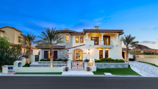 Two-story Mediterranean-style home with stone accents, arched entryways, palm trees, and a broad driveway at dusk.