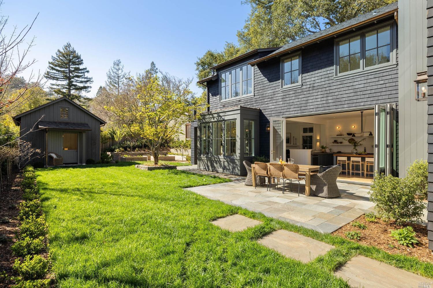 Dark shingle-sided home opening onto a stone patio with outdoor dining furniture and a green lawn.