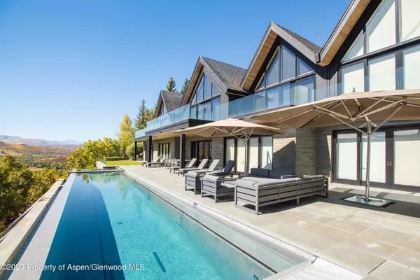 Modern mountain home beside a long pool and furnished patio, with autumn trees and distant mountains.