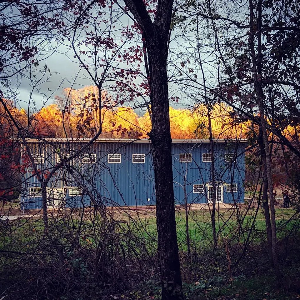 Blue metal-sided building behind bare trees, with autumn foliage on a ridge beyond.