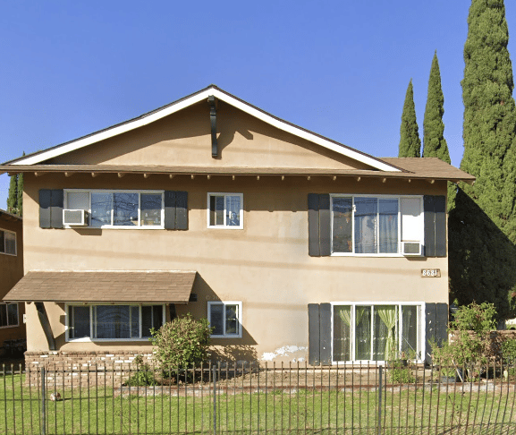 Two-story beige house with dark shutters, window air conditioners, and a fenced front lawn.