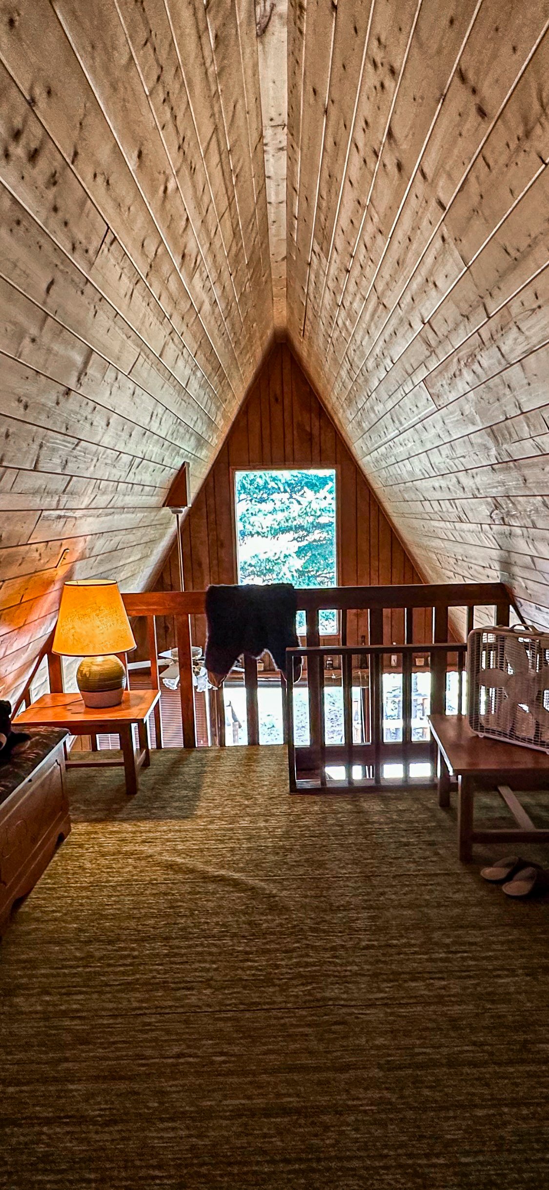 Furnished A-frame loft with vaulted wood ceiling, dark railing, and tall window overlooking trees.