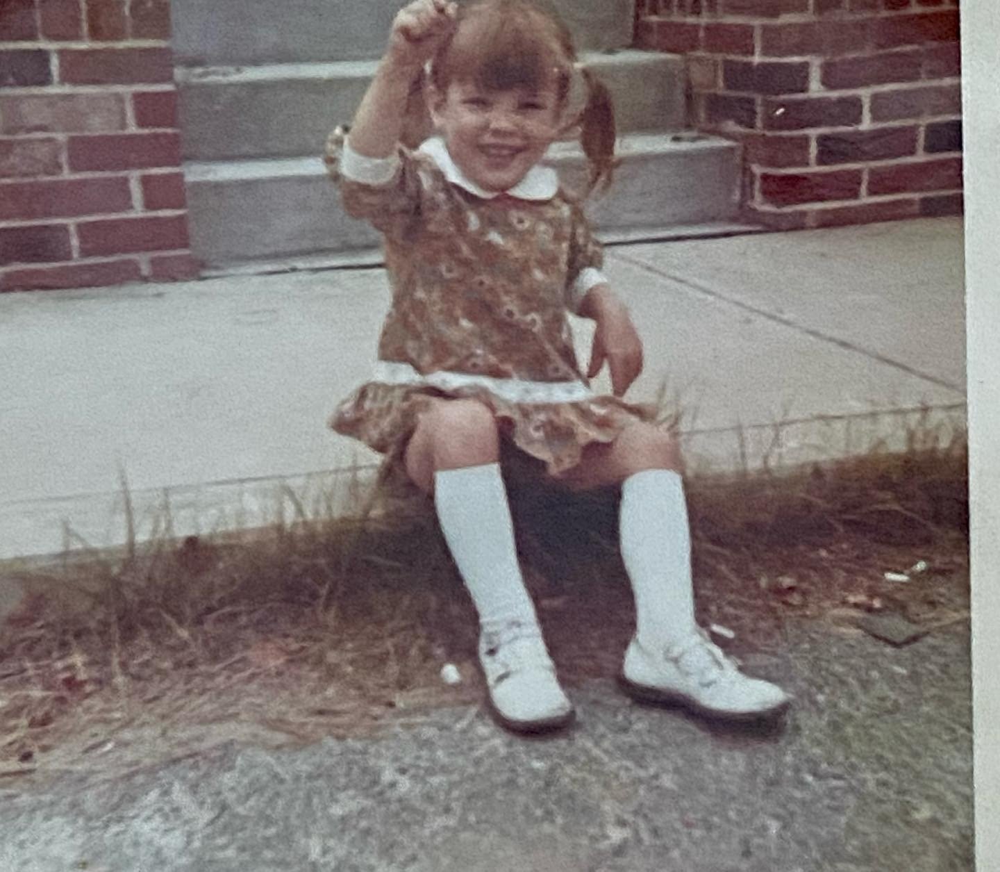 Young girl in a patterned dress sits outside a brick building, smiling and raising one hand.