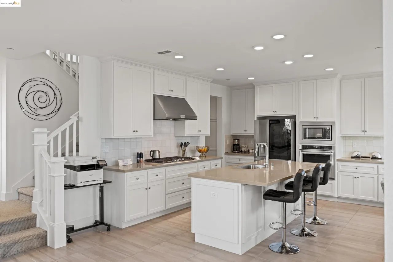 Bright white kitchen with a large island, three bar stools, stainless-steel appliances, and a staircase.