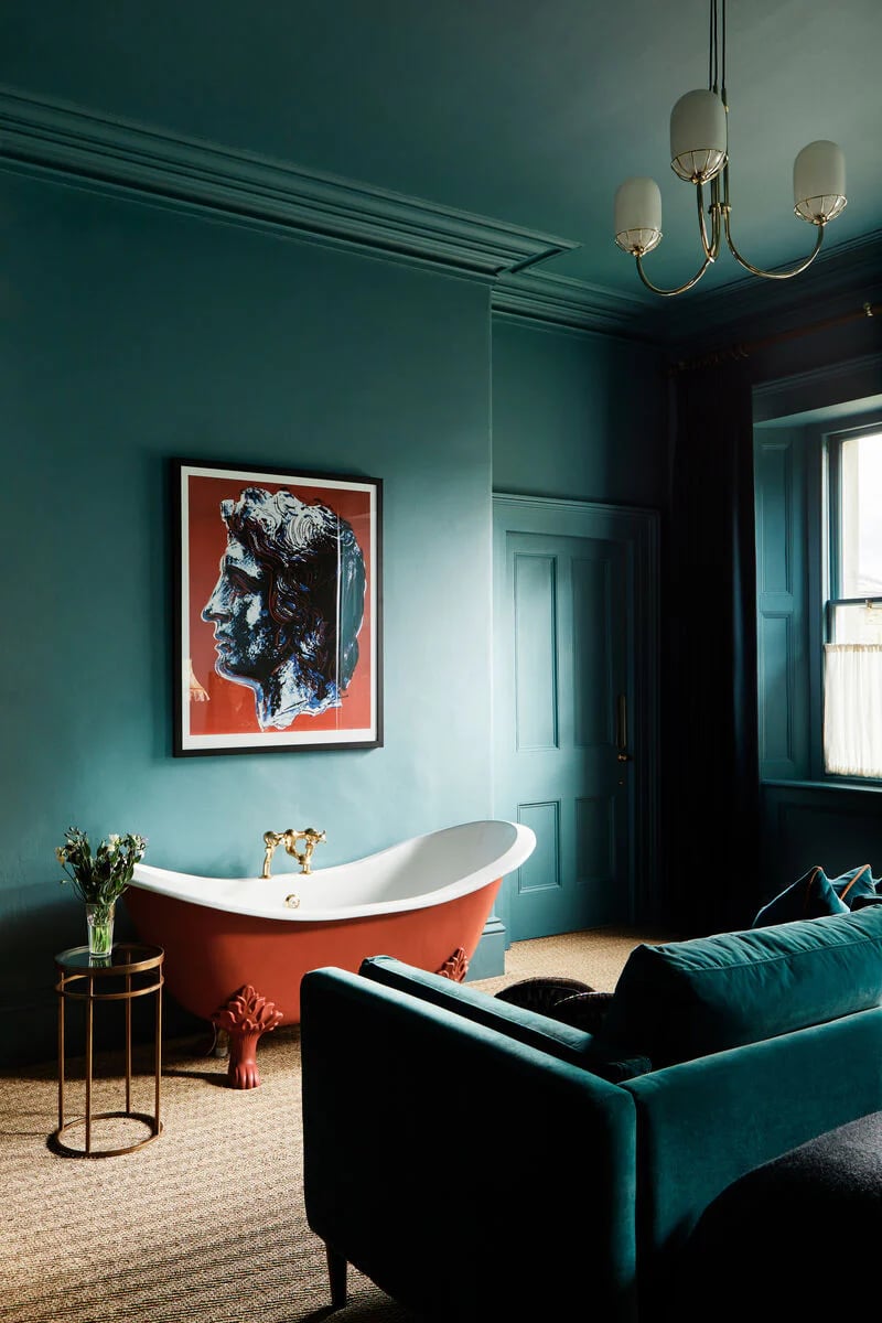 Teal bathroom with a coral clawfoot tub, framed portrait artwork, and a matching velvet sofa.